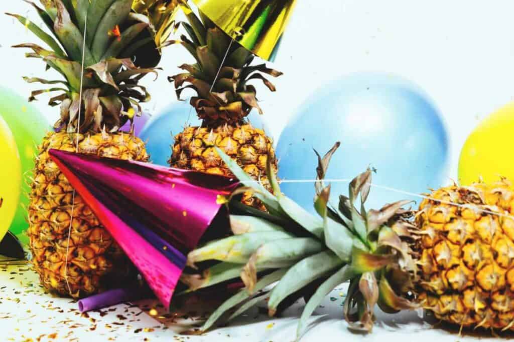 Pineapples, party hats and balloons strewn over a table with confetti.