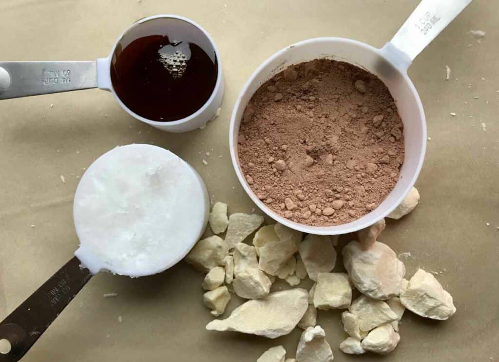 A brown surface with individual measuring cups with cacao powder, coconut oil and maple syrup, beside scattered chunks of cacao butter.
