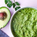 Bowl of guacamole on pink background with half an avocado and a sprig of coriander.