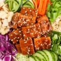 A grain bowl comprised of tofu cooked in a peanut sauce and served with brown rice, cabbage, carrots, cucumber and slivered almonds.