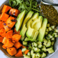 A grain bowl with quinoa or rice, roast sweet potato, red onion, cucumber, avocado, rocket, and pistachio pesto.
