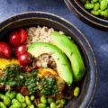 A bowl with caramelised plantains topped with chimichurri sauce on a bed of brown rice alongside edamame beans, cherry tomatoes and avocado.
