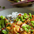 A Japanese-inspired grain bowl combines spicy sriracha tofu with rice, edamame beans and salad greens topped with sesame seeds.