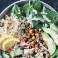A grain bowl with quinoa, crispy garlic herb roasted chickpeas, cucumber, tomato, red onion, avocado, spinach and hummus dressing.