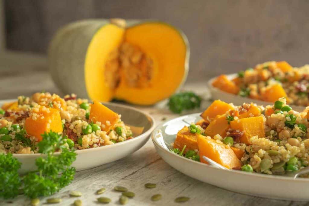 Three green and white plates with pumpkin quinoa salad served on them, sitting on a wooden kitchen bench with a sprinkling of pumpkin seeds, a few sprigs of parsley and pumpkin with a pieced cut out of it in the background.