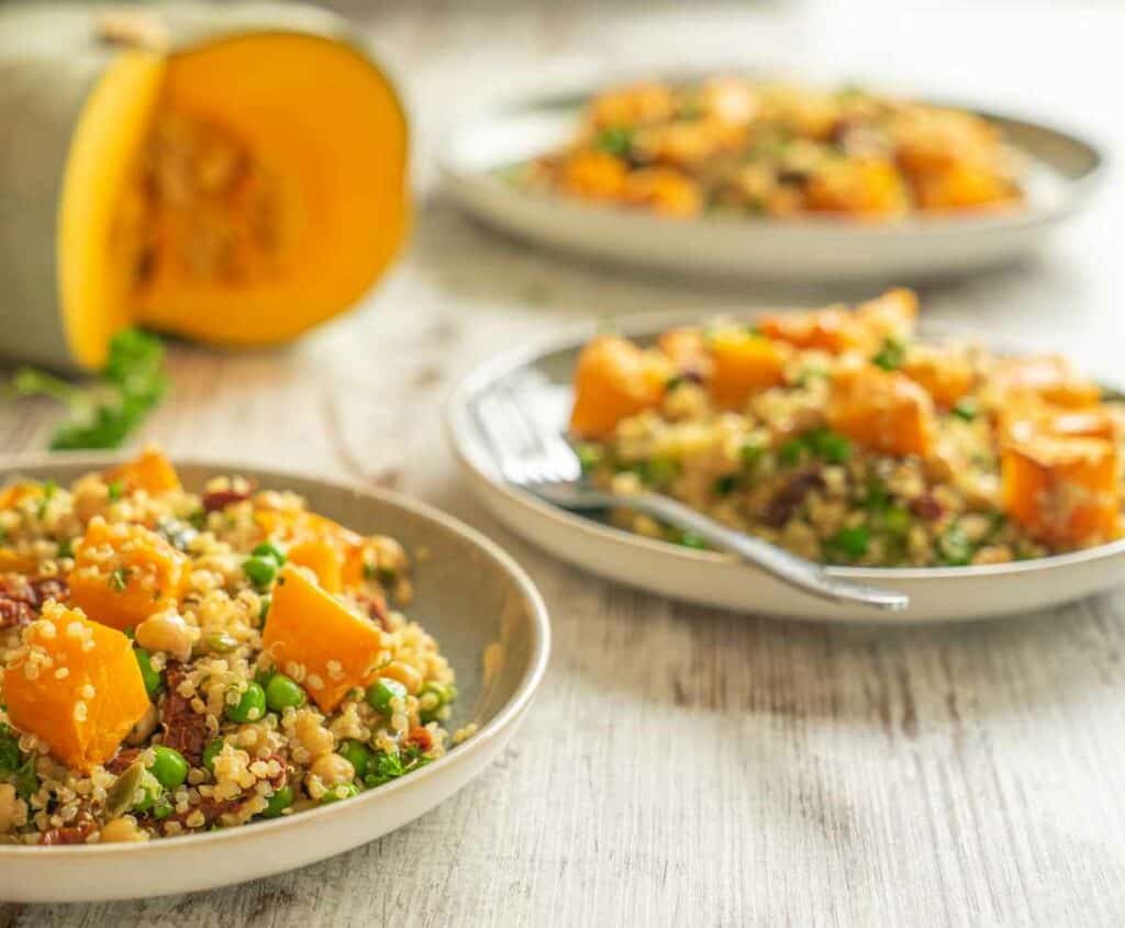 Three green and white plates with pumpkin quinoa salad, sitting on a wooden kitchen bench with a sprig of parsley and pumpkin with a pieced cut out of it in the background.