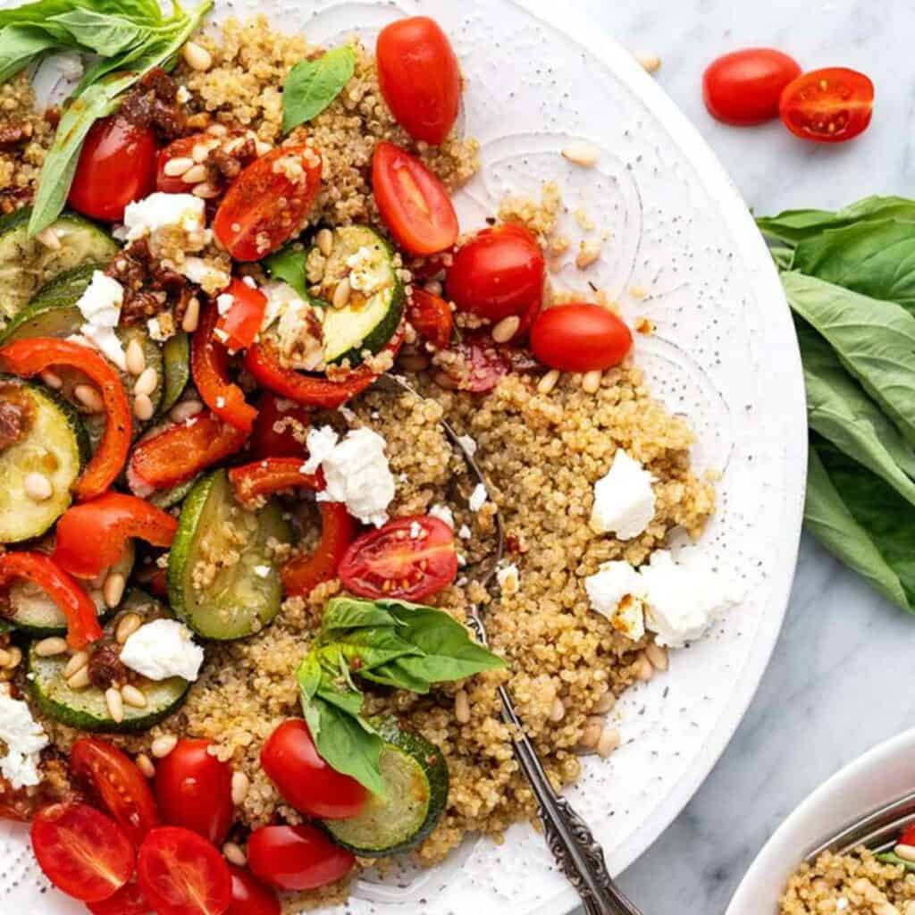 A quinoa salad on a white plate containing roast zucchini, capsicum and tomatoes, topped with a sundried tomato vinaigrette, feta and pine nuts.