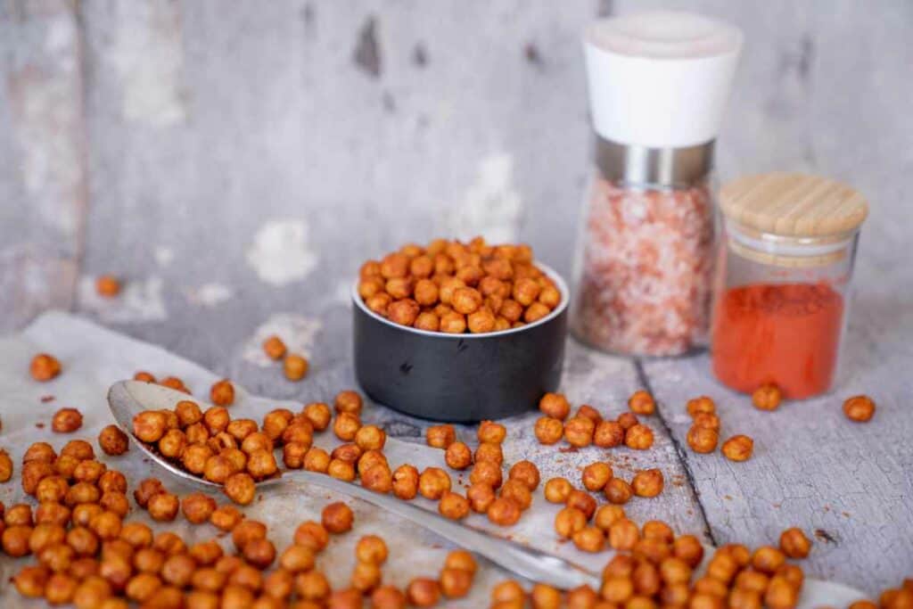 Crispy chickpeas on baking paper with a black bowl of crispy chickpeas and a jar of paprika and salt grinder in background.