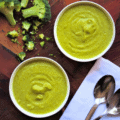 Two small bowls of Vegan Curried Broccoli Soup on wooden chopping bowl with white napkin, two spoons and florets of broccoli.