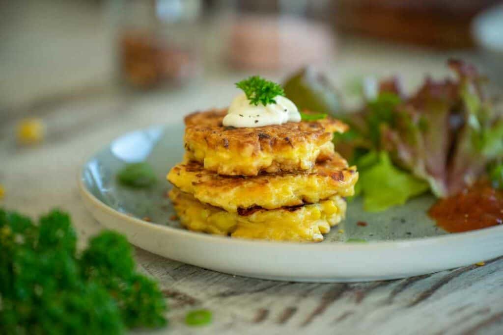 Stack of three corn fritters on a plate with lettuce, relish and sour cream.