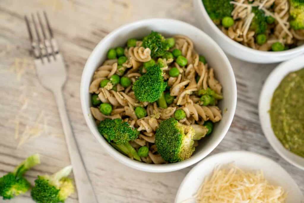 Bowl with vegetarian pesto pasta surrounded by Parmesan cheese, pesto and broccoli florets.
