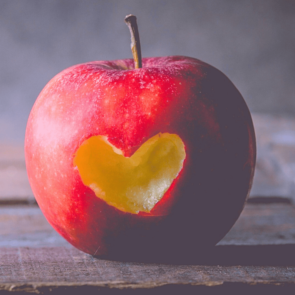 A red apple with a heart shape cut out of it, sitting on a wooden surface.