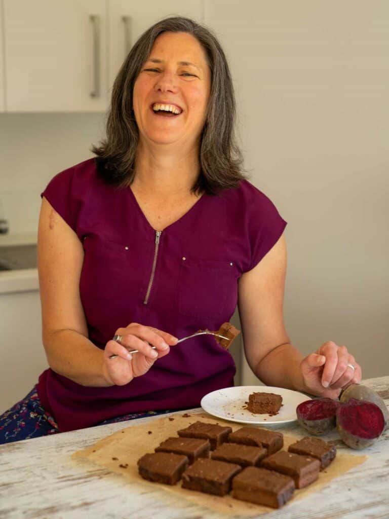 Nutritionist Tansy Boggon laughing, holding a fork, with a plate with a slice of chocolate beetroot brownie on it. In front of the plate are slices of brownie and fresh beetroot.