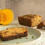 A slice of pumpkin prune loaf on a light green plate with sprinkled pumpkin seeds. In the background is the full loaf and two other slices of the loaf, beside a pumpkin with a quarter cut out of it.