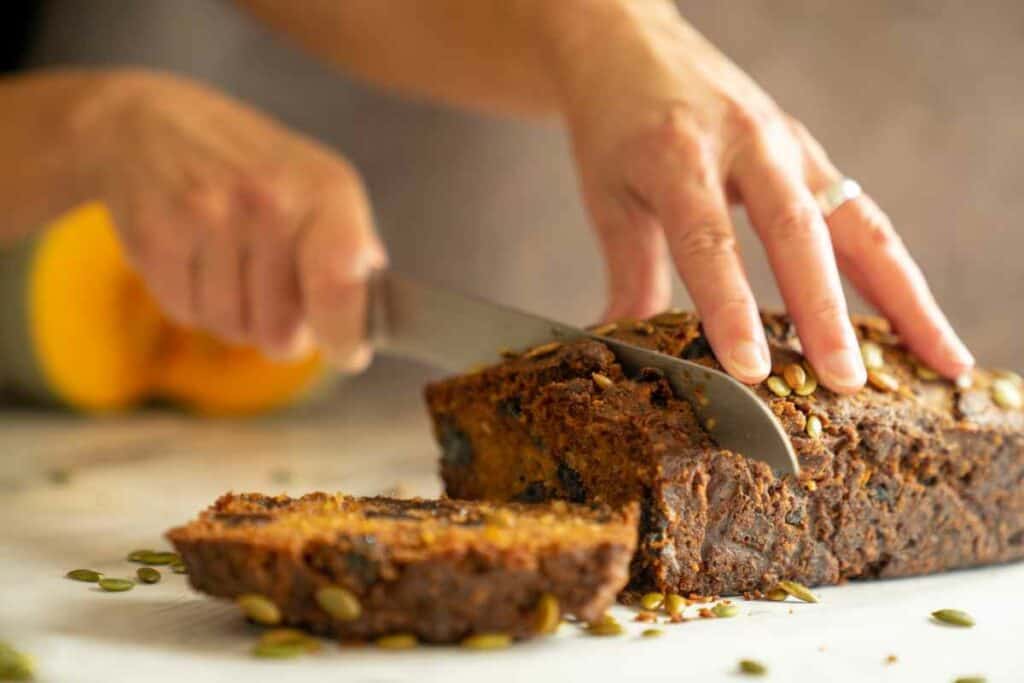 A loaf of pumpkin and prune cake being sliced with a bread knife, with a cut pumpkin in the background.