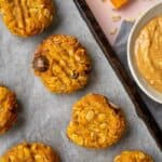 Pumpkin cookies on a black baking tray lined with baking paper, sitting on a light pink surface with a grey tea towel and bowl of peanut butter.