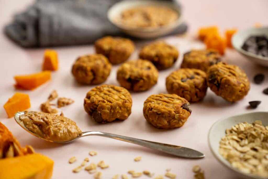 Peanut butter pumpkin cookies on a light pink surface surrounded by a bowls of peanut butter, oats and chocolate chips. On the surface are a a few pieces of cooked pumpkin, oats, chocolate chips and a tablespoon of peanut butter.