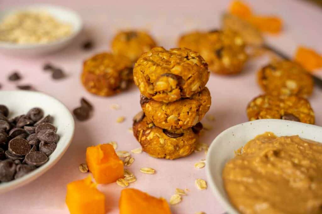 A stack of cookies on a light pink surface surrounded by a bowls of peanut butter, oats and chocolate chips. On the surface are a a few pieces of cooked pumpkin, oats and chocolate chips.
